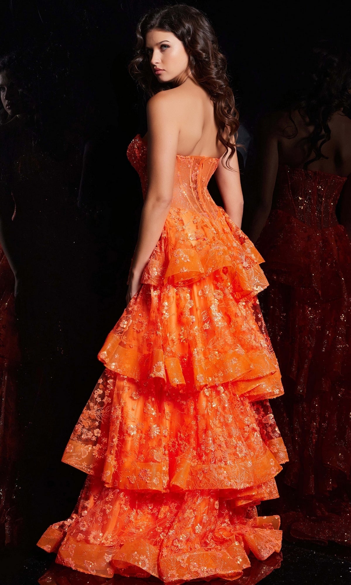 A woman with long, wavy hair wears the Jovani Strapless Long Ruffled Prom Dress 38528 in orange with floral embroidery, standing against a dark background.